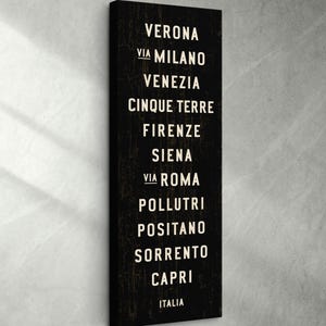 Pu&ograve; includere: Un quadro verticale rettangolare su tela nera con un elenco di citt&agrave; italiane in lettere maiuscole bianche senza grazie. Il testo recita: VERONA, VIA MILANO, VENEZIA, CINQUE TERRE, FIRENZE, SIENA, VIA ROMA, POLLUTRI, POSITANO, SORRENTO, CAPRI, ITALIA.