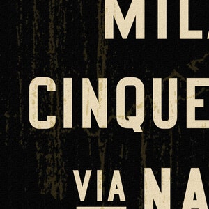 Pu&ograve; includere: Un cartello rettangolare nero con superficie testurizzata che presenta grandi lettere maiuscole sans-serif color bianco sporco. Il testo riporta MIL, CINQUE e VIA NA, con la parola VIA sottolineata. Il design ha un aspetto usurato.