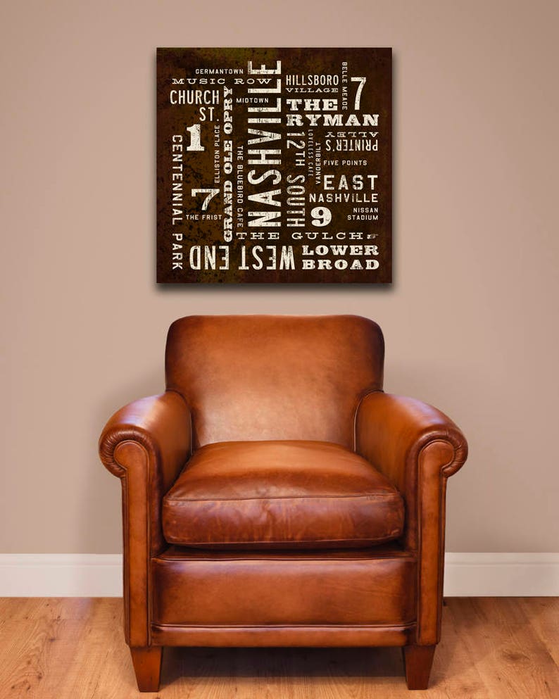 A brown canvas print by Transit Design, with white text that reads 'Nashville' in large letters. The text is surrounded by smaller words that are all related to Nashville, such as 'Music Row', 'Ryman', 'The Gulch', and 'Centennial Park'. The print is hanging on a wall.