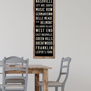 May include: A black and white sign with a wooden frame lists various neighborhoods in Nashville, Tennessee. The text reads: "Nashville, 12th Ave. South, Music Row, Germantown, Belle Meade, Via Belmont, Hillsboro Village, West End, East Nashville, Green Hills, Brentwood, Franklin, Leiper's Fork."