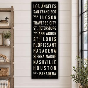 May include: A tall, rectangular black wooden sign featuring white, sans-serif text listing the cities: Los Angeles, San Francisco, via Tucson, Traverse City, St. Petersburg, via Ann Arbor, St. Louis, Florissant, Pasadena, Sierra Madre, Nashville, Houston, so. Pasadena.