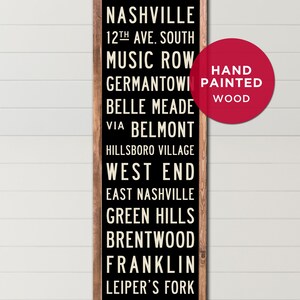 May include: Black wooden sign with white text listing various neighborhoods in Nashville, Tennessee. The sign reads "Nashville", "12th Ave. South", "Music Row", "Germantown", "Belle Meade", "Via Belmont", "Hillsboro Village", "West End", "East Nashville", "Green Hills", "Brentwood", "Franklin", and "Leiper's Fork". A red circle with white text reads "Hand Painted Wood".