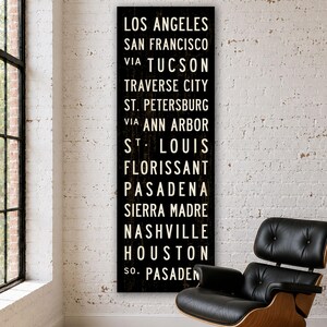 May include: A tall, rectangular black wall sign featuring white, sans-serif text listing the cities: Los Angeles, San Francisco, via Tucson, Traverse City, St. Petersburg, via Ann Arbor, St. Louis, Florissant, Pasadena, Sierra Madre, Nashville, Houston, So. Pasadena.