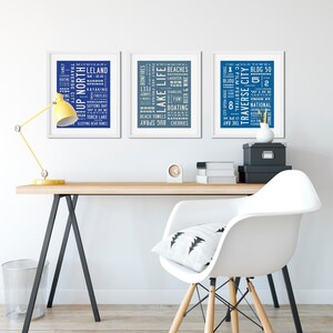 May include: Three framed prints with blue backgrounds and white text. The prints feature word clouds related to the Traverse City, Michigan area. The prints are hanging on a white wall above a wooden desk with a white chair.