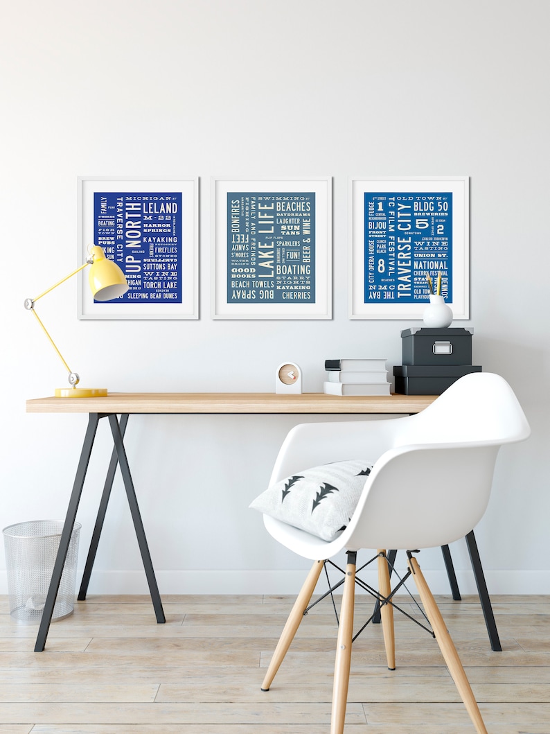May include: Three framed prints with blue backgrounds and white text. The prints feature word clouds related to the Traverse City, Michigan area. The prints are hanging on a white wall above a wooden desk with a white chair.
