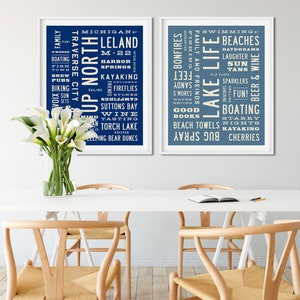 May include: Two blue and white framed prints with white text on a blue background. The left print features the words "Up North" and "Michigan" with a list of activities and places. The right print features the words "Lake Life" and "Swimming" with a list of activities and places.