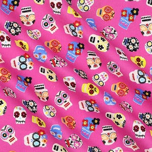 May include: A vibrant pink fabric featuring a repeating pattern of colorful sugar skull designs. The skulls are decorated with floral and geometric patterns in shades of yellow, blue, orange, and black.