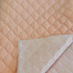 May include: A close-up of a quilted fabric in a soft peach color. The fabric has a diamond pattern created by stitched lines. A section of the fabric is folded over, revealing a lighter, textured underside with the same diamond pattern.