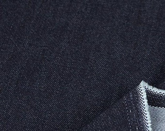 May include: Close-up of a dark blue denim fabric. The fabric has a textured appearance with visible diagonal weave. The edge of the fabric is folded over, revealing a lighter, contrasting edge.