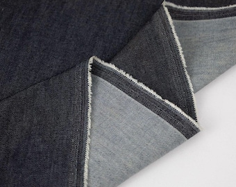 May include: Close-up of dark denim fabric with visible weave texture. The fabric is folded, revealing a lighter gray underside and a raw, frayed edge. The denim has a deep indigo color.