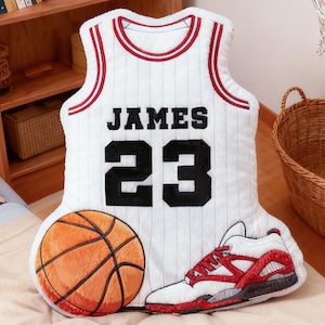 May include: A plush basketball-themed pillow set. The set includes a white jersey pillow with "JAMES 23" in black, a basketball, and a red and white sneaker. The items are arranged on a bed.