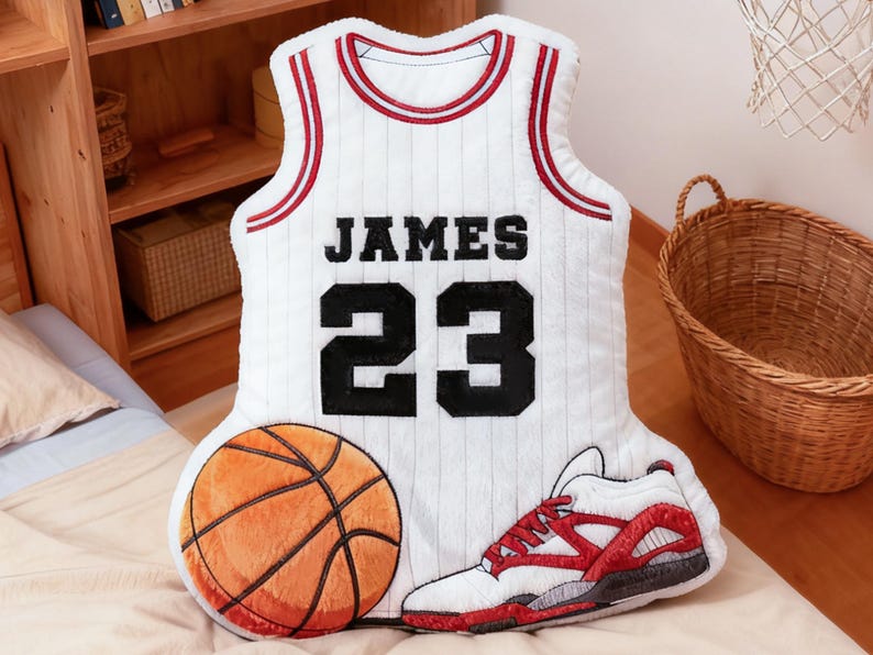 May include: A plush basketball-themed pillow set. The set includes a white jersey pillow with "JAMES 23" in black, a basketball, and a red and white sneaker. The items are arranged on a bed.