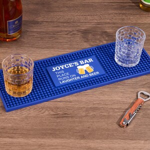 以下が含まれることがあります： 「JOYCE'S BAR」と「THIS PLACE RUNS ON LAUGHTER AND BEER」の文字が入った青いバーマット。琥珀色の液体が入ったグラスが2つと空のグラスがマットの上にあります。栓抜きが近くにあります。