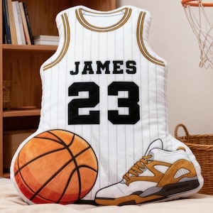 May include: A basketball-themed pillow set featuring a white jersey with "JAMES 23" in black, a basketball, and a sneaker. The jersey has gold trim and vertical stripes. The basketball is orange and brown.