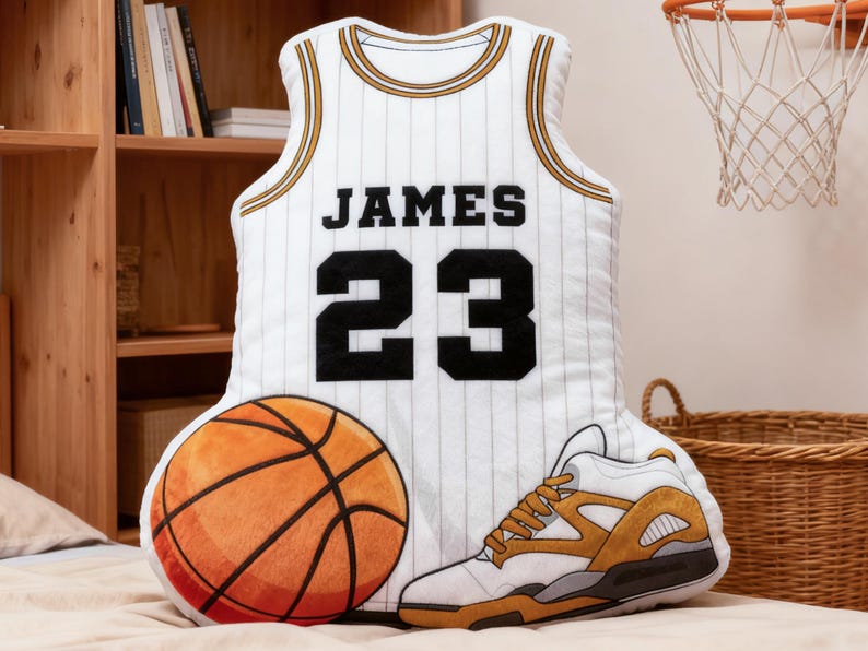 May include: A basketball-themed pillow set featuring a white jersey with "JAMES 23" in black, a basketball, and a sneaker. The jersey has gold trim and vertical stripes. The basketball is orange and brown.
