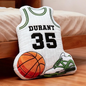 May include: A decorative pillow shaped like a basketball jersey with the name "DURANT" and the number "35" in black. An orange and brown basketball and a white and green sneaker are also featured. The pillow is on a wooden floor.