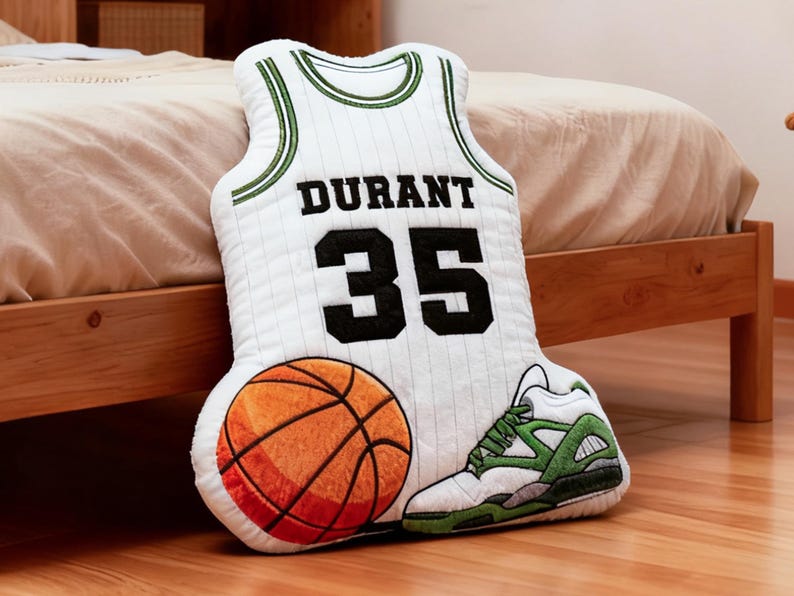 May include: A decorative pillow shaped like a basketball jersey with the name "DURANT" and the number "35" in black. An orange and brown basketball and a white and green sneaker are also featured. The pillow is on a wooden floor.