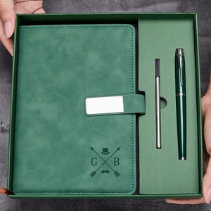 Puede incluir: Un juego de diario y bolígrafo verde en una caja de regalo. El diario tiene una correa con una placa plateada y un diseño con flechas, un sombrero y las letras G y B. El juego incluye un bolígrafo y una recarga.