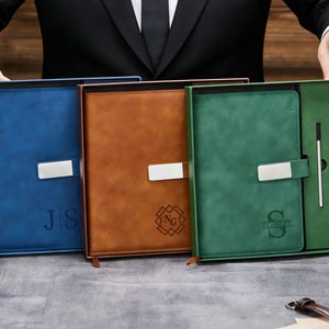 Puede incluir: Tres juegos de diario y bolígrafo encuadernados en cuero azul, marrón y verde. Cada diario tiene un cierre plateado e iniciales o monograma personalizados. El juego verde incluye un bolígrafo y un lápiz. Los artículos se presentan en una caja de regalo.