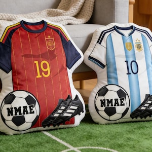 Puede incluir: Dos cojines decorativos con forma de camiseta de fútbol. Uno es rojo con el número 19, el otro es blanco y celeste con el número 10. Cada cojín tiene un balón de fútbol y un par de botas de fútbol negras. El balón de fútbol tiene el texto "NMAE".