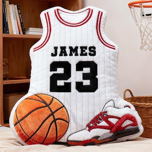 May include: A plush basketball-themed pillow set featuring a white jersey with "JAMES 23" in black, a basketball, and a sneaker. The jersey has red and black accents. The basketball is orange and brown.