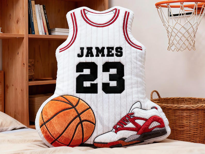 May include: A plush basketball-themed pillow set featuring a white jersey with "JAMES 23" in black, a basketball, and a sneaker. The jersey has red and black accents. The basketball is orange and brown.