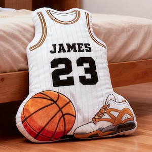 May include: A decorative pillow set featuring a basketball jersey, basketball, and sneaker. The jersey is white with black lettering that reads "JAMES 23". The basketball is orange, and the sneaker is white and orange.
