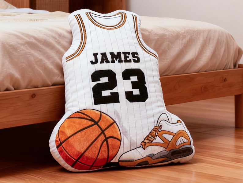 May include: A decorative pillow set featuring a basketball jersey, basketball, and sneaker. The jersey is white with black lettering that reads "JAMES 23". The basketball is orange, and the sneaker is white and orange.