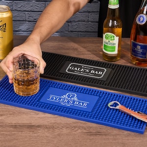 Può includere: Una scena da bar con una lattina dorata di birra Crown Krone, un bicchiere di liquido ambrato e bottiglie di sidro e liquore. Sono visibili due tappetini da bar, uno nero e uno blu. Il tappetino blu ha il testo "Tyler's Bar".