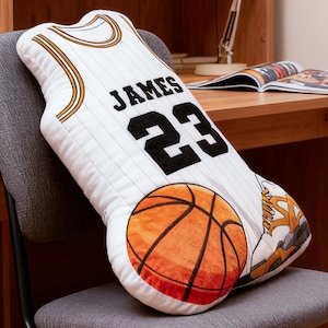 May include: White basketball jersey-shaped pillow with the name "JAMES" and the number "23" in black. The pillow features a basketball and a sneaker design. The pillow is on a gray chair.