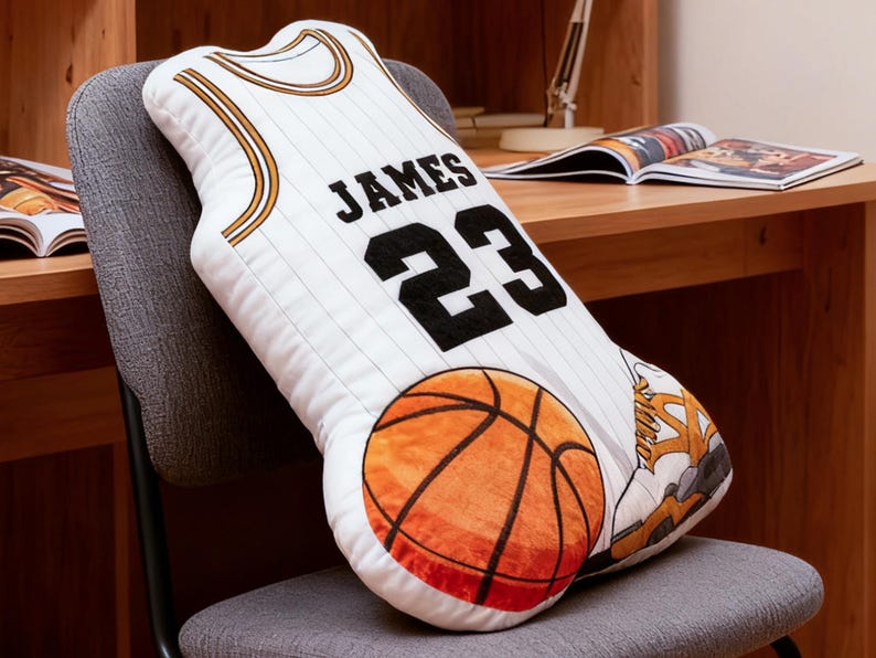 May include: White basketball jersey-shaped pillow with the name "JAMES" and the number "23" in black. The pillow features a basketball and a sneaker design. The pillow is on a gray chair.