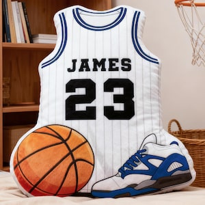 May include: A basketball-themed decorative pillow set featuring a white jersey with "JAMES 23" printed on it, a basketball, and a sneaker. The jersey has blue trim and pinstripes. The basketball is orange and brown.