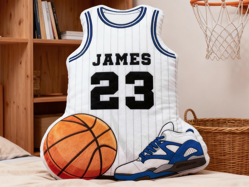 May include: A basketball-themed decorative pillow set featuring a white jersey with "JAMES 23" printed on it, a basketball, and a sneaker. The jersey has blue trim and pinstripes. The basketball is orange and brown.