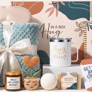 May include: A gift set featuring a light blue textured blanket with a white ribbon, a white mug with the text Hug in a Mug, a notebook labeled Get Better Every Day, a candle, a crochet heart plant, a bath bomb, soap, and socks.