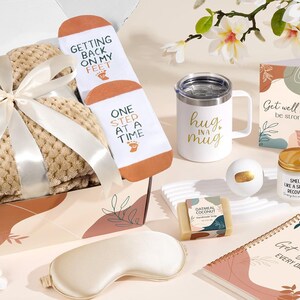 May include: A gift box containing a beige waffle-knit blanket tied with a white ribbon, white socks with the text Getting Back On My Feet One Step At A Time, a white mug with Hug In A Mug, a Get Well Soon card, a candle, soap, a notebook, and a crochet heart.