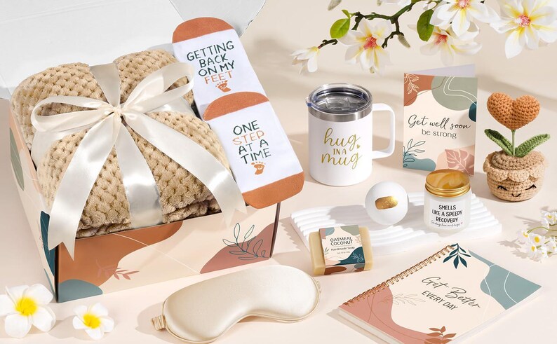 May include: A gift box containing a beige waffle-knit blanket tied with a white ribbon, white socks with the text Getting Back On My Feet One Step At A Time, a white mug with Hug In A Mug, a Get Well Soon card, a candle, soap, a notebook, and a crochet heart.