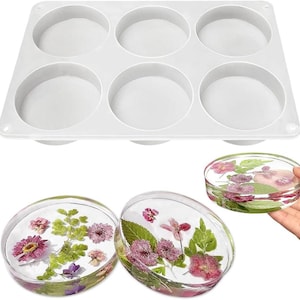 May include: A white silicone mold with six circular cavities, alongside three clear resin coasters embedded with dried flowers. The coasters showcase pink, purple, and green floral arrangements, offering a glimpse of the finished product.
