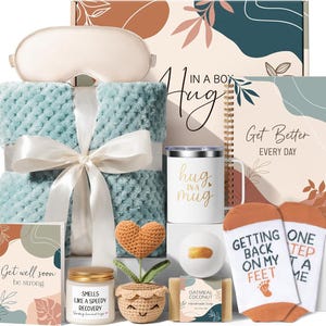 May include: A gift box with a variety of items, including a blue blanket, eye mask, mug, socks, and a notebook. The box is decorated with floral designs and the words "Hug in a Box". Other items include a candle, soap, and a crocheted heart.