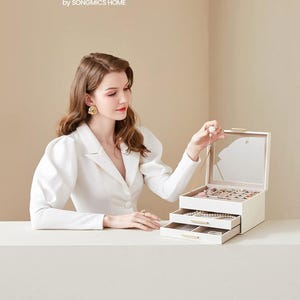 May include: A cream-coloured jewellery box with three drawers and a mirrored lid. The box is open, displaying necklaces, bracelets, and rings. The brand name "SONGMICS" is visible in the top left corner.