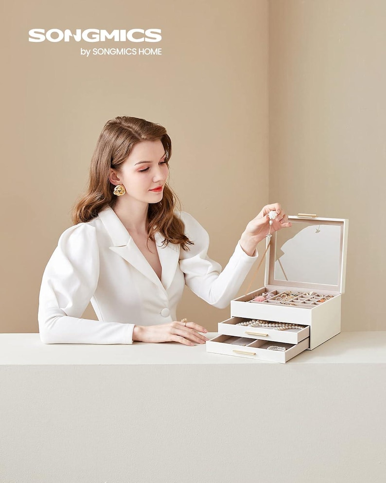 May include: A cream-coloured jewellery box with three drawers and a mirrored lid. The box is open, displaying necklaces, bracelets, and rings. The brand name "SONGMICS" is visible in the top left corner.