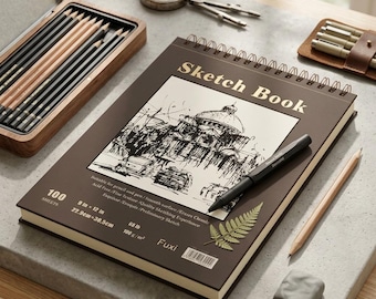 May include: A brown spiral-bound sketchbook with the text Sketch Book on the cover, featuring a black and white architectural drawing, a black pen, a fern leaf, and printed specifications including 9 in x 12 in and 100 sheets.
