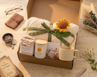 May include: A gift box containing a cream-colored cable knit blanket tied with a green ribbon, a crocheted sunflower, a pair of beige fuzzy socks, a white candle with a wooden lid, a greeting card, and a ribbed ceramic mug with a gold spoon.