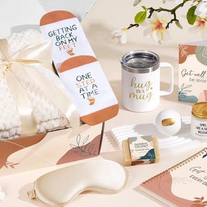 May include: A gift box containing a white textured blanket with a cream ribbon, white socks with the text Getting Back On My Feet One Step At A Time, a white mug with Hug In A Mug, a Get Well Soon card, a candle, soap, a bath bomb, and a notebook.