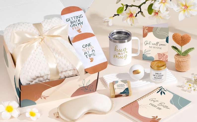 May include: A gift box containing a white textured blanket with a cream ribbon, white socks with the text Getting Back On My Feet One Step At A Time, a white mug with Hug In A Mug, a Get Well Soon card, a candle, soap, a bath bomb, and a notebook.