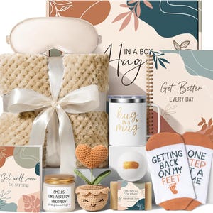 May include: A gift set containing a beige textured blanket with a white ribbon, a sleep mask, a white mug with the text 'hug in a mug', a notebook labeled 'Get Better EVERY DAY', a crochet heart plant, a candle, soap, and socks with the text 'GETTING BACK ON MY FEET ONE STEP AT A TIME'.
