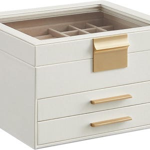 May include: A white jewellery box with a clear glass top and gold-coloured hardware. The box has a top compartment with dividers, two drawers, and a gold-coloured handle. The box is designed for storing jewellery.