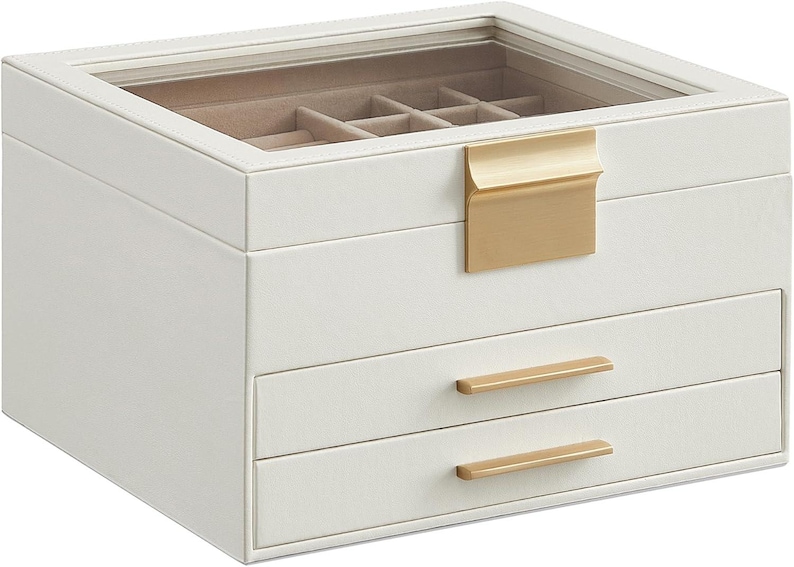 May include: A white jewellery box with a clear glass top and gold-coloured hardware. The box has a top compartment with dividers, two drawers, and a gold-coloured handle. The box is designed for storing jewellery.