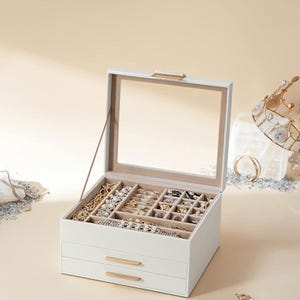 May include: A white jewelry box with a clear glass top and two drawers. The box is filled with various necklaces, bracelets, rings, and earrings. Gold-toned hardware accents the drawers and top. A crown and jewelry are visible in the background.