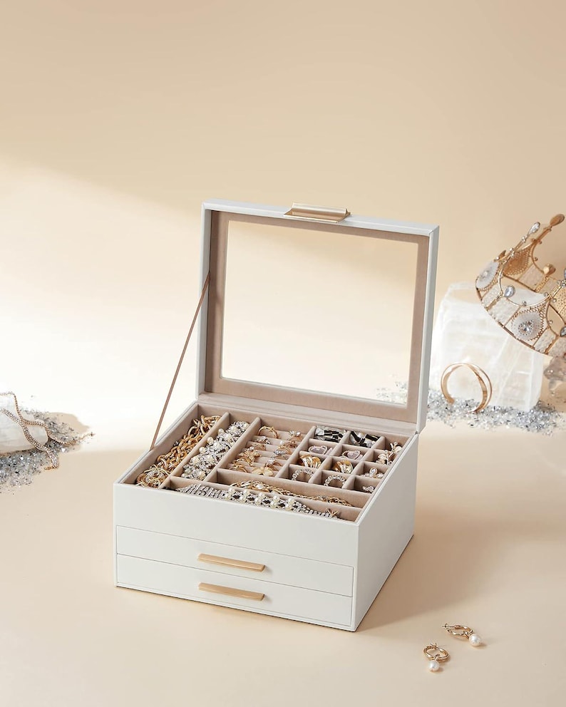 May include: A white jewellery box with a clear glass top and two drawers. The box is filled with various necklaces, bracelets, rings, and earrings. Gold-toned hardware accents the drawers and top. A crown and jewellery are visible in the background.