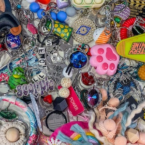 May include: A selection of keyrings in various colours and designs. Includes a pink paw print, a green frog, a pop-it toy, and charms with the words "Scorpio" and "Power". There are also charms with the text "Hollywood Production" and "Battery Shine".